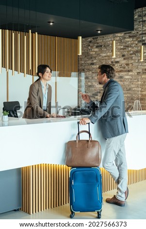 Female Hotel Receptionist Assisting Businessman for Checking In.

Business man with luggage talking with concierge on hotel reception with sneeze guard protection.