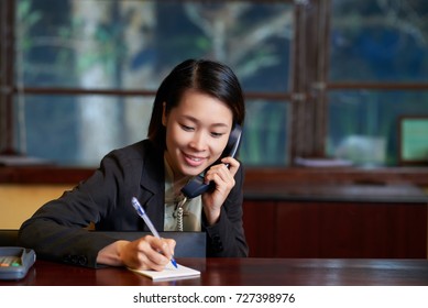 Female Hotel Receptionist Talking On The Phone And Taking Notes