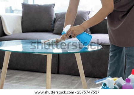 Female housekeeper wearing apron and casual clothes standing at table with buckets of cleaning equipments holding alcohol spray and cloth to clean and wipe glass table in living room at home.