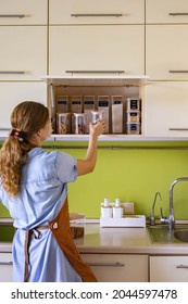 Female Housewife Placing Glass Jars Full Of Healthy Food Nuts, Grains, Cereals Assortment At Cupboard Shelf For Comfortable Storage. Woman Neatly Putting Dry Products Keeping Organization At Cuisine