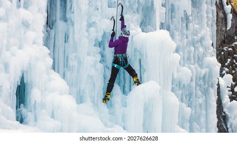 Female ice climber with ice climbing equipment, axes, helmet, harness, and crampons hanging on a frozen waterfall, back view