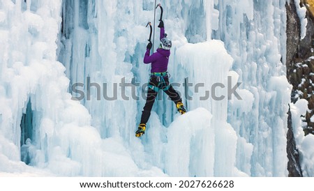 Female ice climber with ice climbing equipment, axes, helmet, harness, and crampons hanging on a frozen waterfall, back view