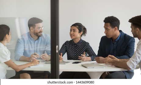Female indian company representative talking at diverse group briefing sit at conference table behind glass door, business woman manager team leader consult clients colleagues negotiating in office