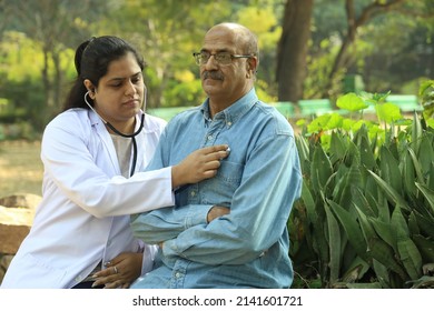 Female Indian Doctor Examining The Senior Patient Who Is Holding A Walking Stick In The City Park Afternoon. 