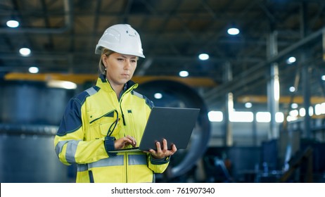 Female Industrial Engineer in the Hard Hat Uses Laptop Computer while Standing in the Heavy Industry Manufacturing Factory. In the Background Various Metalwork Project Parts Lying