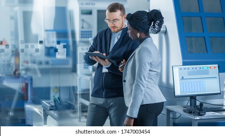 Female Industrial Engineer Stands With Male Project Supervisor, They Use Tablet Computer And Discuss Modern Factory Planning. Manufacturing Facility With CNC Machinery And Robot Arm Assembly Line