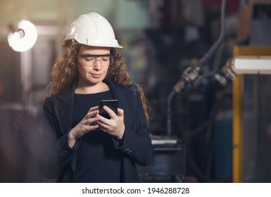 Female Industrial Worker In The Hard Hat Uses Mobile Phone While Walking Through Heavy Industry Manufacturing Factory. 