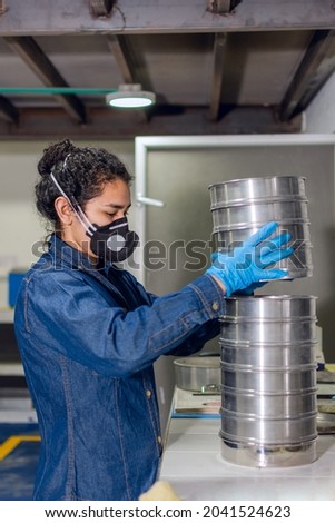Female industrial worker with protective mask and blue latex gloves, organizing metal containers in a geological testing laboratory