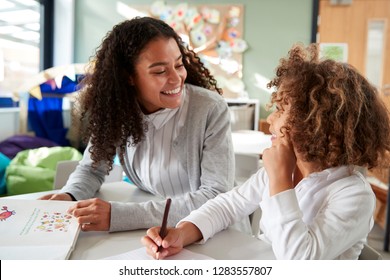 Female infant school teacher working one on one with a young schoolgirl, sitting at a table smiling at each other, front view, close up