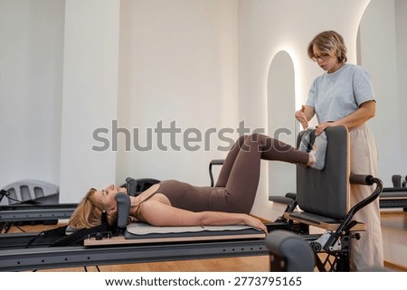 Female instructor guiding client through leg extension routine on pilates reformer in airy gym. Leg extension, pilates reformer, instructor, routine, gym