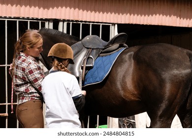 Female instructor guiding young girl in saddling a horse. Young rider wearing safety helmet and polo shirt outdoor - Powered by Shutterstock