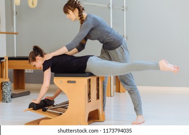 Female instructor of pilates helping to young woman workout on chair working out indoor in pilates studio, correcting beginners, full height. Treatment of back pain concept. - Shutterstock ID 1173794035