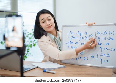 Female instructor presents an interactive math lesson, solving equations on a whiteboard while engaging with students through a laptop camera in a bright, modern classroom.
