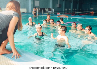 Female Instructor Teaches Children How To Swim. Kids With Goggles In Water Listening Trainer. Happy Kids In Modern Sport Center. Concept Of Fun, Leisure And Recreation.