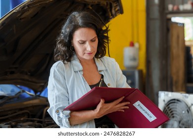 Female insurance agent reviewing documents inside auto repair shop. Woman holding folder labeled "Insurance", focused on paperwork.  administrative tasks, customer service, and vehicle insura - Powered by Shutterstock