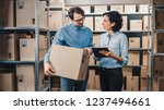 Female Inventory Manager Shows Digital Tablet Information to a Worker Holding Cardboard Box, They Talk and Do Work. In the Background Stock of Parcels with Products Ready for Shipment.