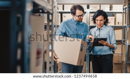 Female Inventory Manager Shows Digital Tablet Information to a Worker Holding Cardboard Box, They Talk and Do Work. In the Background Stock of Parcels with Products Ready for Shipment.