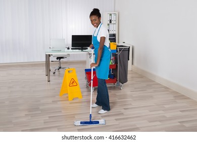 Female Janitor Mopping Wooden Floor With Caution Sign In Office