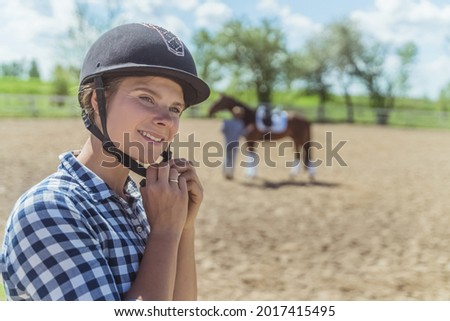 Female jockey putting on a helmet and getting ready for horse riding. A girl with a dark bay horse in the background. Sandy parkour with a wooden fence. Horse riding for leisure.