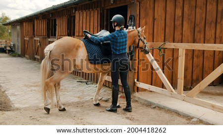Female jockey saddling up her light brown horse outside the stable. Placing a leather saddle on the back of her horse. The girl wearing a helmet and gloves. Preparing her horse for the competition.