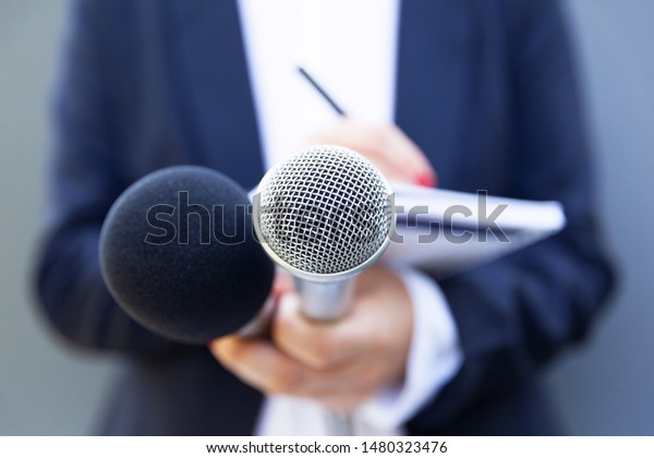 Female journalist at news conference\
or media event, writing notes, holding\
microphone