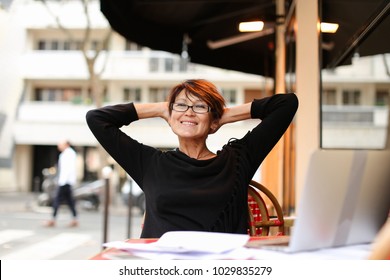 Female Journalist Working Hard For Long Time At Laptop For Important Report Article. Middle Aged Lady Dressed In Black Blouse Have Short Haircut And Wear Glasses. Woman Finishing Printing In Computer