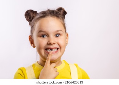 Female kid with opened mouth pointing at missing front baby tooth with finger smiling excitedly in yellow t-shirt on white background. First teeth changing. Going to dentist to do tooth treatment. 