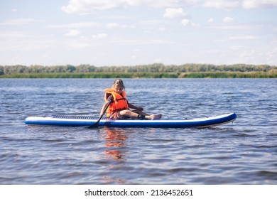 Female kid sitting on sup board by herself on lake holding oar in right hand and looking at rippled water in orange life jacket. Active holidays. Inculcation of love for sports from childhood. - Shutterstock ID 2136452651