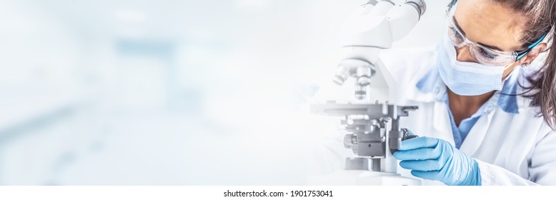 Female lab technician in protective glasses, gloves and face mask sits next to a microscope and conical flask, looking aside on the desk - panoramic banner. - Powered by Shutterstock