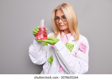 Female laboratory worker observes chemical processes focused at glass flask with red liquid wears medical coat and round spectacles isolated over grey background. Woman researcher conducts experiment - Powered by Shutterstock