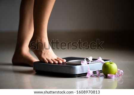Female leg stepping on weigh scales with measuring tape and green apple. Healthy lifestyle, food and sport concept.