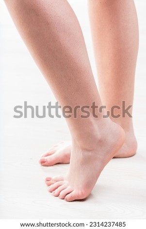 Female legs, bare feet with pedicured, polished nails. Left leg, foot in foreground, right slightly blurred behind. Background is light wood floor. Fair skin. Personal care, beauty, wellness