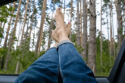 Female Legs In Blue Jeans Out Of Car Window. Concept Of Comfortable Local Travel Vacation Holiday. Reduce Carbon Footprint. Woman Resting In Road Trip 