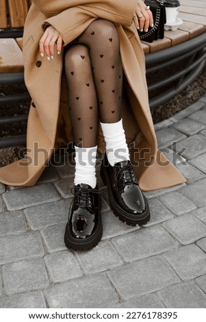 female legs dressed in heart print tights, white socks, black patent leather massive shoes brogues, lifestyle model dresses in brown oversized coat, details of stylish fashion outfit