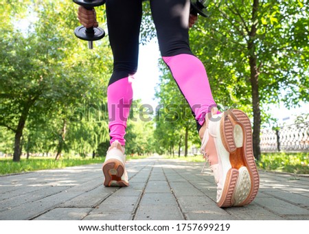 Female legs and feet in leggings and sneakers. Faceless woman does step forward with dumbbells in public park outdoor in summer, wide angle, back view, selective focus. Training, workout, fitness