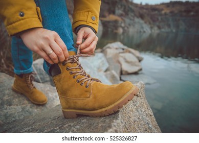 Female Legs In Jeans And Hiking Boots Near Edge Of Mountain Lake. Rocks Reflection On Water Surface