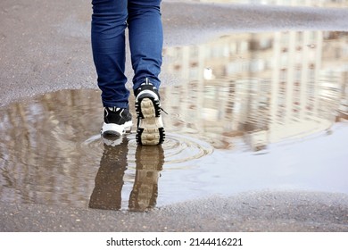 Female legs in jeans and sneakers with reflection in puddle on a street. Rain in city, melting snow in spring,  footwear for woman