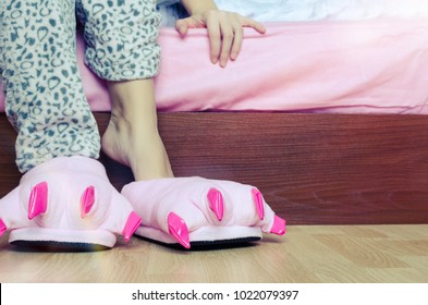 Female Legs In Warm Pajama And Cute Pink Monster Foot Slippers With Big Claws On Parquet Floor Near A Bed In Bedroom. Bedtime And Good Morning Concept.