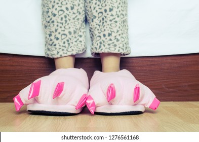 Female Legs In Warm Pajama And Cute Pink Monster Foot Slippers With Big Claws On Parquet Floor Near A Bed In Bedroom. Bedtime And Good Morning Concept.