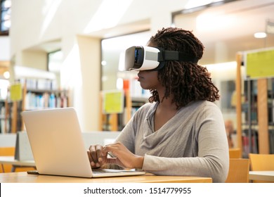 Female library user watching virtual video. Young African American woman wearing virtual reality glasses, sitting at desk with laptop. Innovation for education concept - Powered by Shutterstock