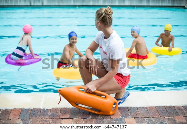 Female lifeguard holding rescue can while children\
swimming in pool