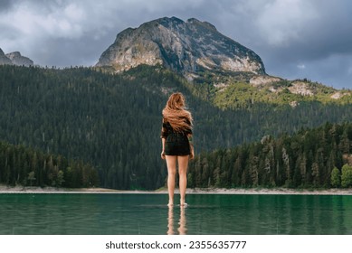 Female with long flying hair standing in water and enjoying view of Black lake in Durmitor, Montenegro. Woman wearing plaid shirt admires panorama of glacial lake surrounded by wild coniferous forest. - Powered by Shutterstock