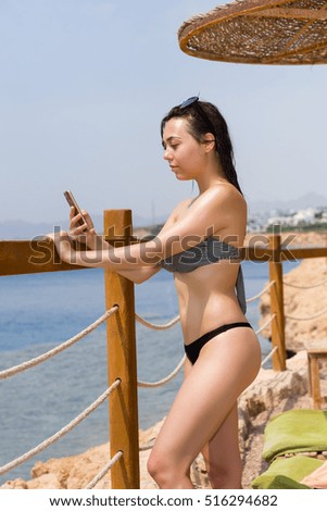 Female is looking for something in the Internet or taking pictures on her phone while standing in front of wooden fence with rope inserts and sea with corals and reefs against sky