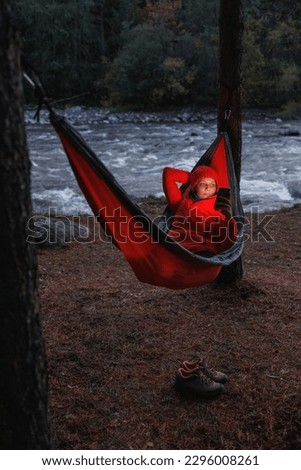 female is lying in hammock and chatting in smartphone, woman in red jacket in nature with flashlight or smartphone shines in dark. camping or picnic in the forest in mountains, solo hiking for person