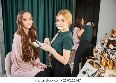 Female Makeup Artist Looking At Camera And Smiling While Client Sitting On Chair In Beauty Salon. Beauty Specialist In Face Mask Holding Eyelashes Set While Standing Near Young Woman In Visage