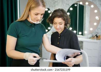 Female makeup artist in protective mask holding pencil and smiling while showing papers to client. Young woman and beauty specialist studying documents in visage studio. - Shutterstock ID 2167231199