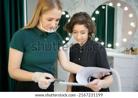 Female makeup artist in protective mask holding pencil and smiling while showing papers to client. Young woman and beauty specialist studying documents in visage studio.