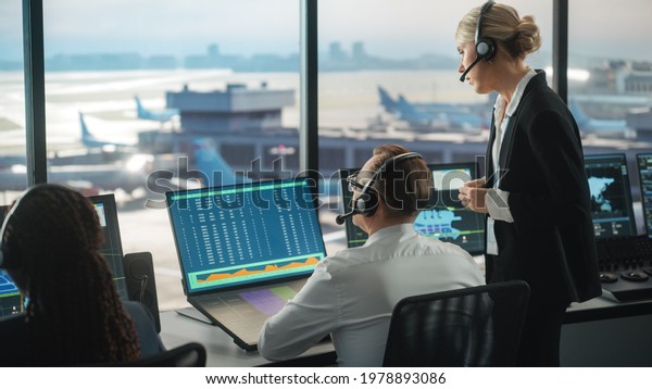 Female and Male Air Traffic Controllers with
Headsets Talk in Airport Tower. Office Room is Full of Desktop
Computer Displays with Navigation Screens, Airplane Flight Radar
Data for the Team.