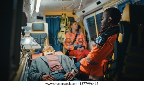 Female and Male EMS Paramedics Ride to Healthcare\
Hospital with an Injured Patient on the Ambulance. Black African\
American Emergency Care Assistantis Sitting Next Sick Young Man\
with Neck Collar.