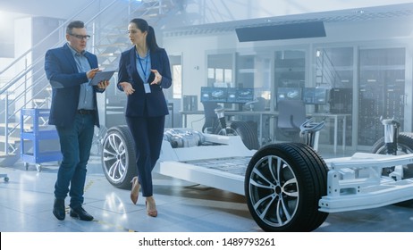 Female And Male Engineer Walk In A High Tech Development Facility Holding A Tablet Computer And Pass An Electric Car Chassis Prototype With Batteries And Wheels.